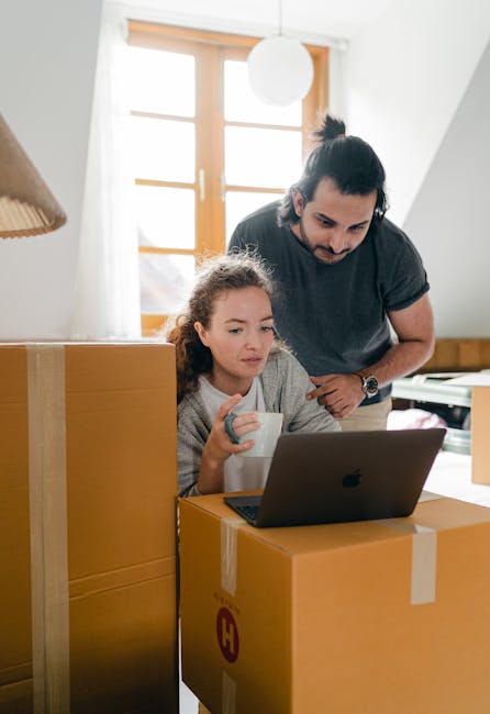 A woman and a man are inside a residential property during a home relocation process. The woman, sitting on a cardboard box, is holding a white mug and working on a laptop placed on top of another box, with a focused expression. The man is standing behind her, leaning slightly forward, observing the screen. Both are in a bright room illuminated by natural light coming through a large window with wooden frames, which is visible in the background. The scene includes several cardboard boxes, some sealed with packing tape, and a few larger pieces of furniture, indicating an active packing and moving environment. The setting captures the logistics involved in furniture transport and packing during a house move, consistent with the services offered by [COMPANY_NAME], which specialises in removals and relocation services in Kingston upon Thames.