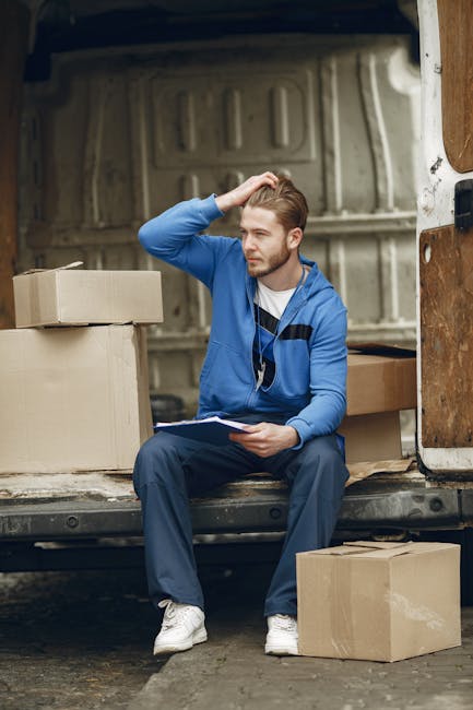 A young man with short, light brown hair and a beard is sitting inside the open back of a van during a home relocation. He is wearing a blue hoodie, dark trousers, and white sneakers, and he has wired earphones around his neck. He appears to be taking a break, holding a clipboard with papers in his left hand and scratching his head with his right hand. Surrounding him are several cardboard boxes, some stacked inside the van and a few placed on the ground nearby, indicating an ongoing packing and loading process. The van's interior is visible, showing a light-colored, slightly textured wall and some wooden panels. The scene suggests a professional removals service, such as that provided by manandvankingstonuponthames.com, organizing or executing a furniture transport and home relocation, with the focus on a careful and methodical approach to moving household items.