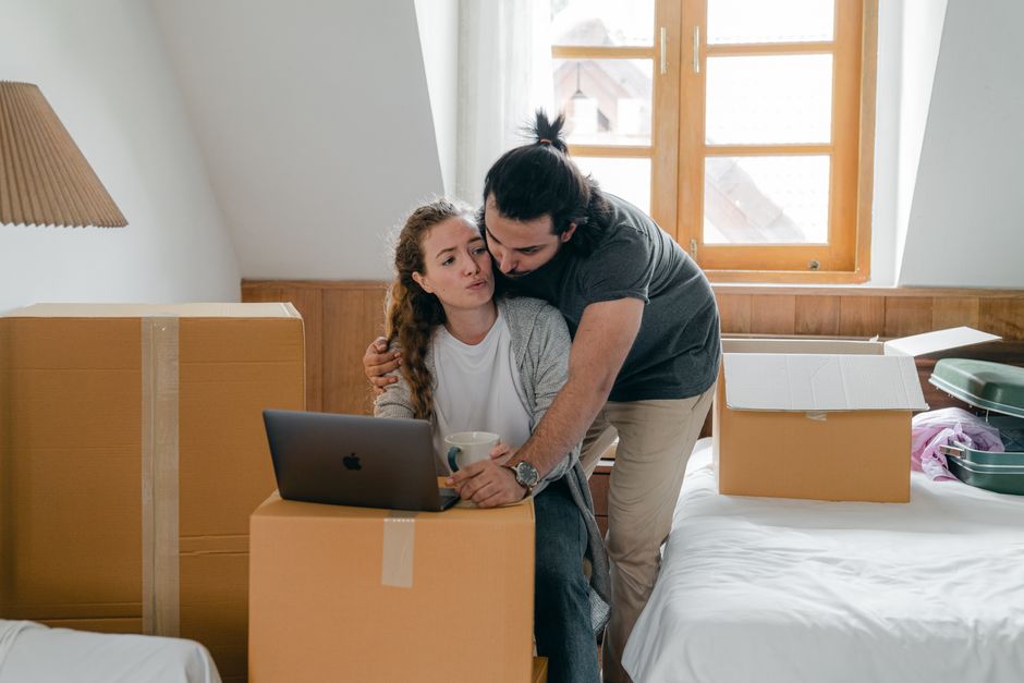 A woman and a man are inside a residential property during a home relocation process. The woman, sitting on a cardboard box, is holding a white mug and working on a laptop placed on top of another box, with a focused expression. The man is standing behind her, leaning slightly forward, observing the screen. Both are in a bright room illuminated by natural light coming through a large window with wooden frames, which is visible in the background. The scene includes several cardboard boxes, some sealed with packing tape, and a few larger pieces of furniture, indicating an active packing and moving environment. The setting captures the logistics involved in furniture transport and packing during a house move, consistent with the services offered by [COMPANY_NAME], which specialises in removals and relocation services in Kingston upon Thames.