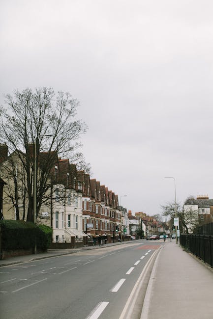 A quiet urban street scene in Kingston upon Thames showing a row of residential buildings with brick facades and pitched roofs on the left side, partially obscured by leafless trees. The street has multiple lanes with white dashed markings and a designated cycling lane on the right, separated by a curb. There are no vehicles or pedestrians visible, and the sky is overcast, giving a neutral light. In the distance, streetlights and a bus stop are visible. This setting may represent an area where house removals or relocations could take place, with the clear roadway suitable for loading a removal van. The image is relevant for illustrating the environment for home mobility services, including packing, furniture transport, and logistics involved in house removals, as handled by local companies such as manandvankingstoneuponthames.com.