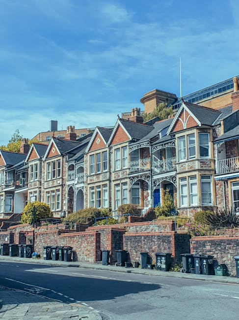 View of a row of Victorian-style residential houses on Tudor Road in Kingston upon Thames, featuring ornate white iron balconies, bay windows, and gabled roofs with red brick and white trim. The properties are elevated above the pavement, separated by brick walls and landscaped garden areas with shrubbery. The street in the foreground has parked black wheelie bins along the curb, and the road appears to be used for vehicle parking and loading. The sky above is partly cloudy with patches of blue, and the scene captures a calm residential neighbourhood suitable for house removals and moving services, such as packing, furniture transport, and home relocation processes. The image is provided by manandvankingstonuponthames.com, illustrating a typical local setting for property moves in Kingston upon Thames.
