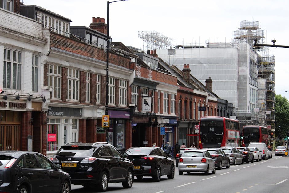 A quiet urban street scene in Kingston upon Thames showing a row of residential buildings with brick facades and pitched roofs on the left side, partially obscured by leafless trees. The street has multiple lanes with white dashed markings and a designated cycling lane on the right, separated by a curb. There are no vehicles or pedestrians visible, and the sky is overcast, giving a neutral light. In the distance, streetlights and a bus stop are visible. This setting may represent an area where house removals or relocations could take place, with the clear roadway suitable for loading a removal van. The image is relevant for illustrating the environment for home mobility services, including packing, furniture transport, and logistics involved in house removals, as handled by local companies such as manandvankingstoneuponthames.com.