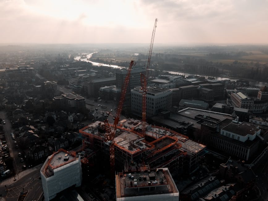 An aerial view of a construction site in Kingston upon Thames shows a multi-storey building in the process of being erected, with several tall orange cranes positioned around the structure. The building's framework is partially completed, with concrete slabs and steel reinforcements visible. Surrounding the site are various commercial and residential buildings, streets with parked cars, and a river flowing in the background, all under overcast skies. The scene demonstrates the ongoing development in the area, which could be related to future housing or commercial space, relevant to home or business relocations. This setting highlights the importance of organized logistics and professional removals services, such as those provided by [COMPANY_NAME], in managing property moves within evolving urban environments.