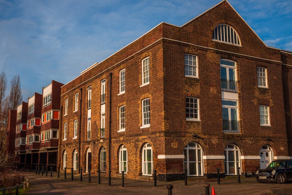 A multi-storey brick Georgian-style building situated on a city street, with large arched windows on the ground floor and smaller rectangular windows on the upper floors, some with black metal balconies. The building's exterior features a mix of red and brown bricks, with white horizontal and vertical accents around the windows and doors. To the left, a row of modern apartment units with protruding balconies in red and white extends along the street. The scene is captured in natural daylight with a partly cloudy sky, and the pavement in front of the building is lined with evenly spaced black bollards. A black car is parked on the street near the building's entrance. This image relates to house removals and home relocation services, illustrating a typical urban residential area where furniture transport and packing are part of the moving process, as provided by [COMPANY_NAME].