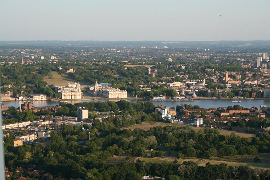 An aerial view of a construction site in Kingston upon Thames shows a multi-storey building in the process of being erected, with several tall orange cranes positioned around the structure. The building's framework is partially completed, with concrete slabs and steel reinforcements visible. Surrounding the site are various commercial and residential buildings, streets with parked cars, and a river flowing in the background, all under overcast skies. The scene demonstrates the ongoing development in the area, which could be related to future housing or commercial space, relevant to home or business relocations. This setting highlights the importance of organized logistics and professional removals services, such as those provided by [COMPANY_NAME], in managing property moves within evolving urban environments.
