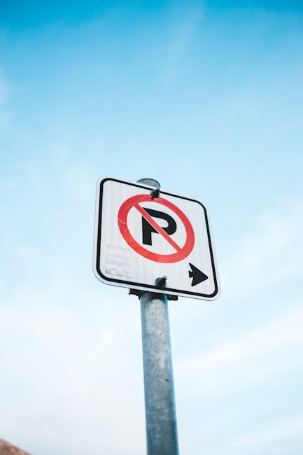 A white rectangular parking restriction sign mounted on a metal pole against a pale blue sky, displaying a black letter 'P' with a red circle and diagonal line through it, indicating no parking. An arrow points to the right beneath the symbol. The sign is situated on a pavement near a property entrance in Kingston upon Thames, and is relevant to local parking regulations for house removals and furniture transport services. The image captures the signage clearly, providing context for parking rules that may impact home relocation or moving logistics in the Kingston KT1 KT2 area, in relation to services offered by manandvankingstonuponthames.com.