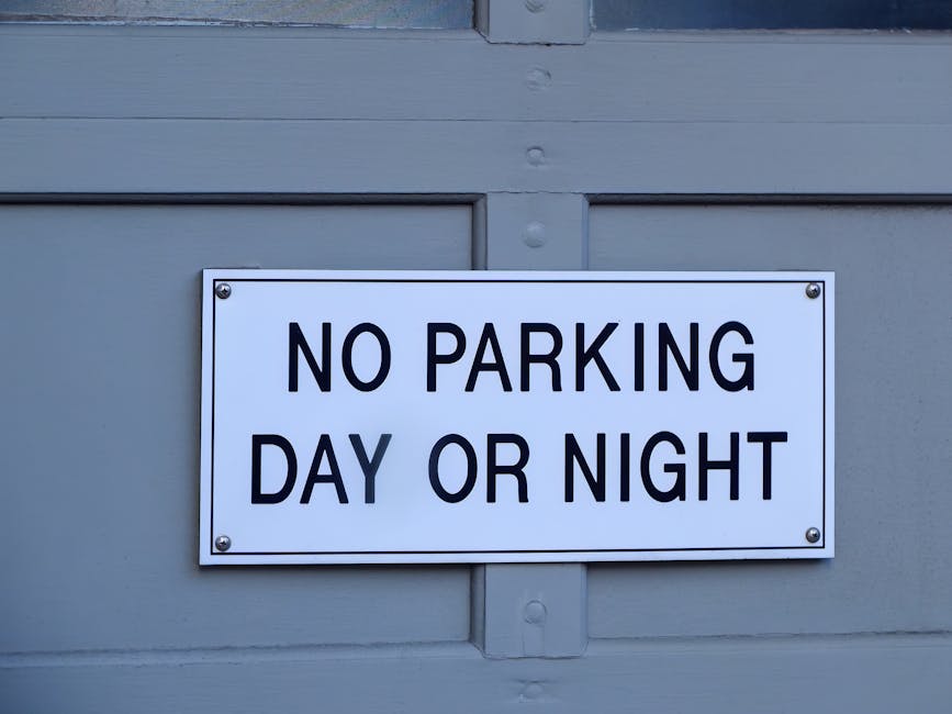 A white rectangular parking restriction sign mounted on a metal pole against a pale blue sky, displaying a black letter 'P' with a red circle and diagonal line through it, indicating no parking. An arrow points to the right beneath the symbol. The sign is situated on a pavement near a property entrance in Kingston upon Thames, and is relevant to local parking regulations for house removals and furniture transport services. The image captures the signage clearly, providing context for parking rules that may impact home relocation or moving logistics in the Kingston KT1 KT2 area, in relation to services offered by manandvankingstonuponthames.com.