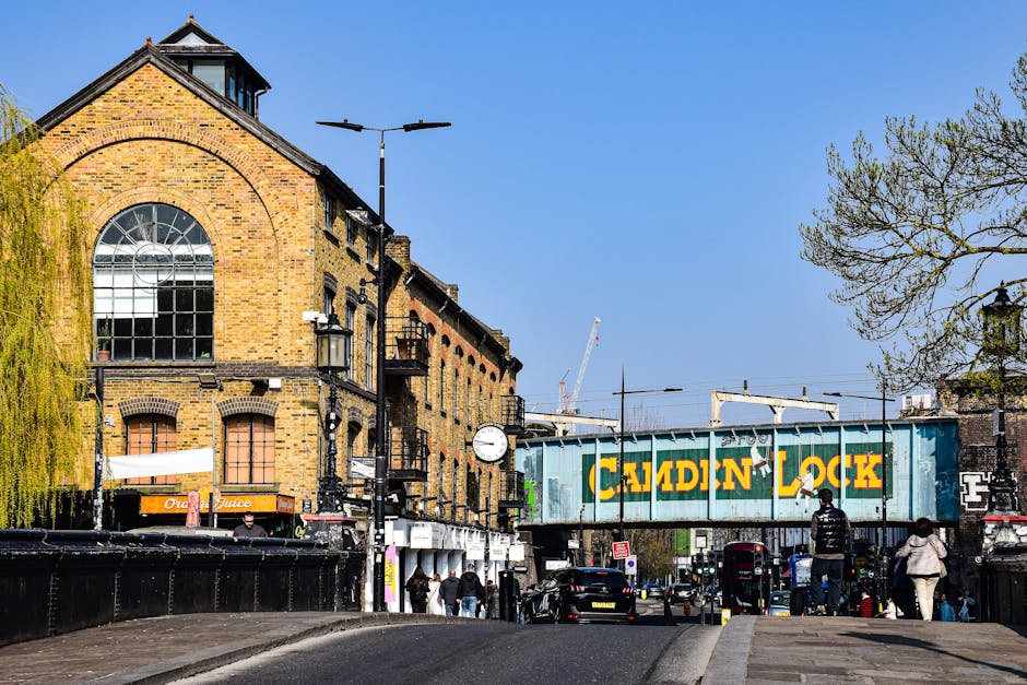 A street scene on Kingston High Street in Kingston upon Thames, featuring a brick building with large arched windows on the left, and a blue railway bridge with yellow and green 'Caldin Lock' signage crossing over the road. Several pedestrians are walking on the sidewalk, some carrying bags, while cars and a red double-decker bus are visible on the road. A black van is parked near the building, and construction cranes can be seen in the background, indicating ongoing development. The scene is well-lit with clear blue sky, and a mixture of trees with budding leaves provides greenery. This setting relates to local house removals and furniture transport services, with the street environment highlighting typical moving logistics in Kingston upon Thames.