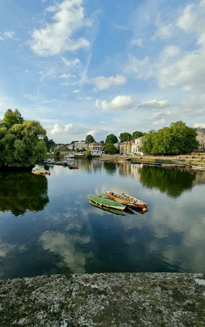 A tranquil view of the River Thames in Kingston upon Thames with several small boats moored along the calm water's edge. The surface of the river reflects the partly cloudy blue sky above, with white fluffy clouds scattered across. On the opposite bank, there are traditional British buildings, some with light-colored facades, and lush green trees lining the promenade. The scene is well-lit with natural daylight, capturing the peaceful atmosphere typical of the Kingston High Street area during daytime. This scenic riverside setting, with visible boats and surrounding architecture, is relevant for visual context related to house removals or relocation services in the Kingston upon Thames area, as shown on the webpage about local removals guidance by manandvankingstonuponthames.com.