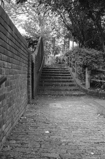 A black-and-white photograph showing a steep outdoor staircase made of concrete or stone, ascending between a brick wall on the left and a landscaped garden area with bushes and trees on the right. The stairs are narrow with visible wear on the treads, and there is a metal handrail on the right side. The surrounding environment appears shaded, with dappled light filtering through the trees above. The scene captures the challenging access point often encountered during house removals or home relocations, illustrating the type of steep stairs that [manandvankingstonuponthames.com](https://manandvankingstonuponthames.com) might address when providing moving services in Kingston upon Thames.